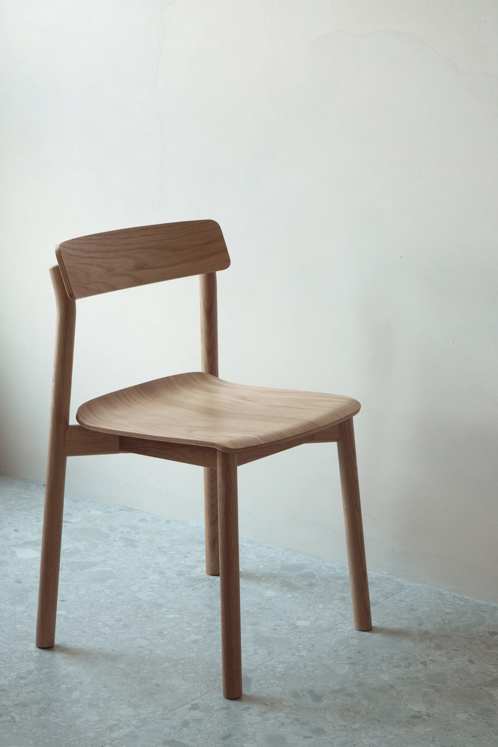 TAMO Arche Chair. A premium, light brown natural oak wood chair with a curved backrest and sculpted seat, displayed against a white wall on gray terrazzo flooring. Perfect for modern dining or office spaces.