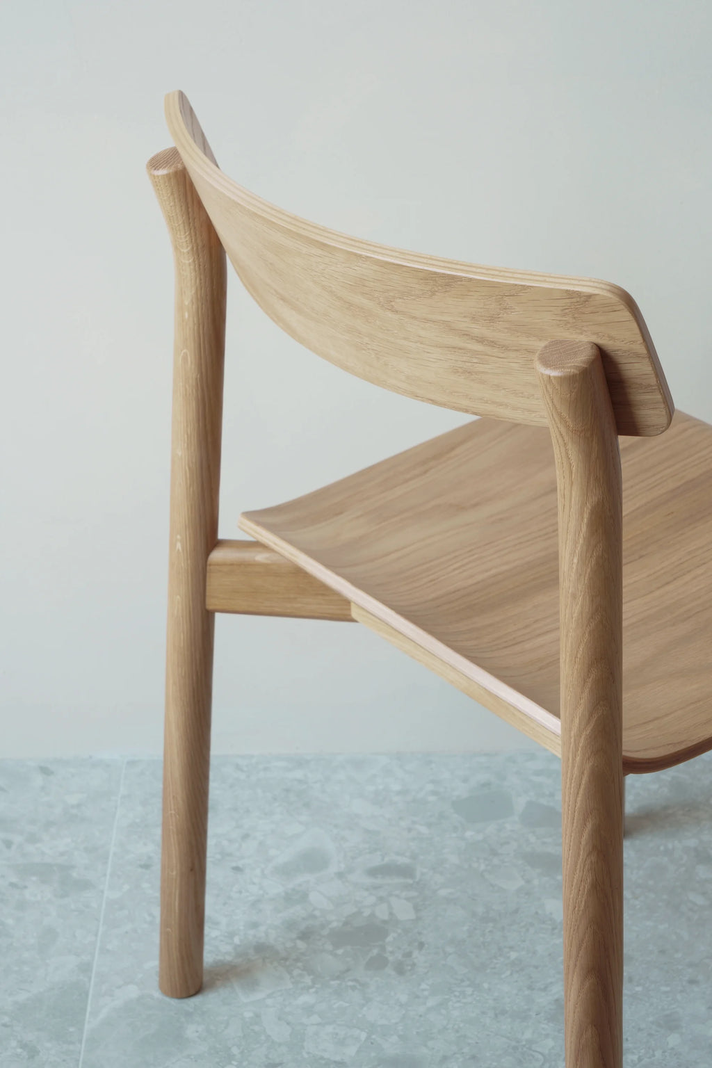 TAMO Arche Chair. A premium light oak wood chair with curved backrest and seat, showcasing elegant minimalist design from a rear-side view on patterned grey flooring. Perfect for modern living spaces.