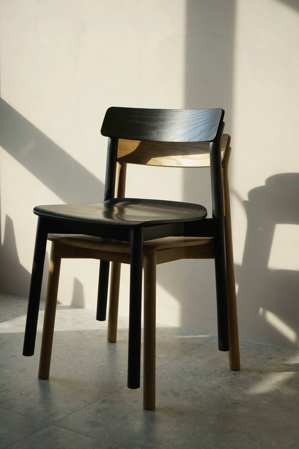 TAMO Arche Chair, a modern chair with a dark wood seat and back, and natural light wood legs, featuring distinctive wood grains, stacked in a sunlit display. Ideal for contemporary dining spaces.