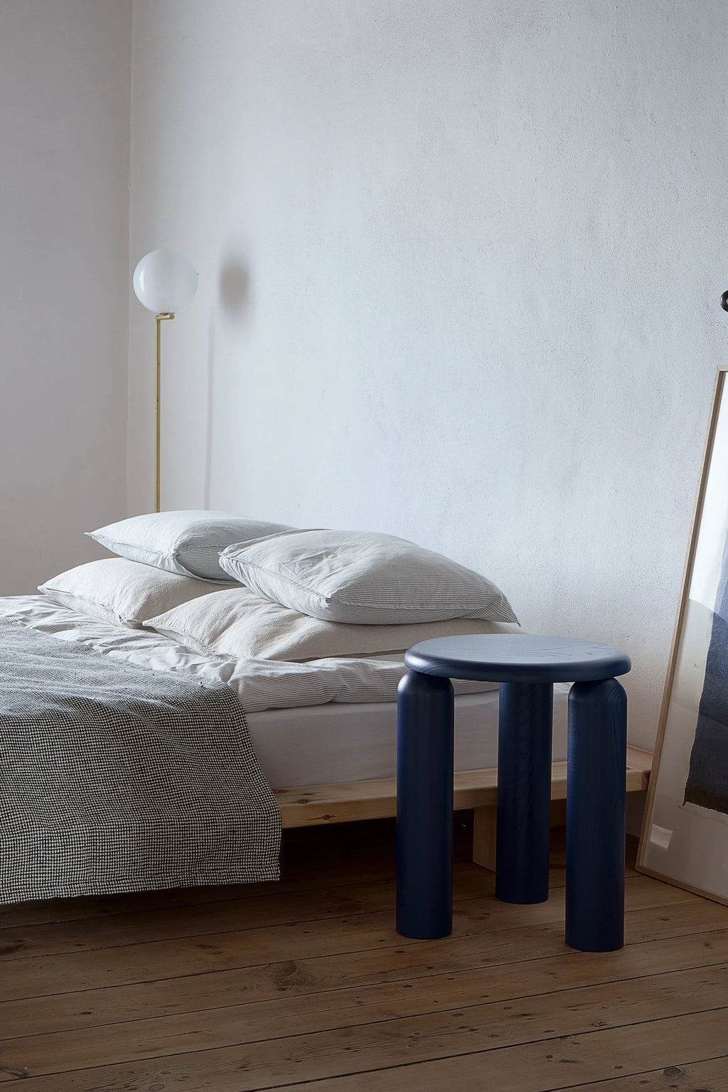 TAMO Neo Side Table in a lifestyle shot. This modern navy-blue wooden side table with three thick, round legs and a smooth top sits beside a bed. A quality piece for contemporary living.