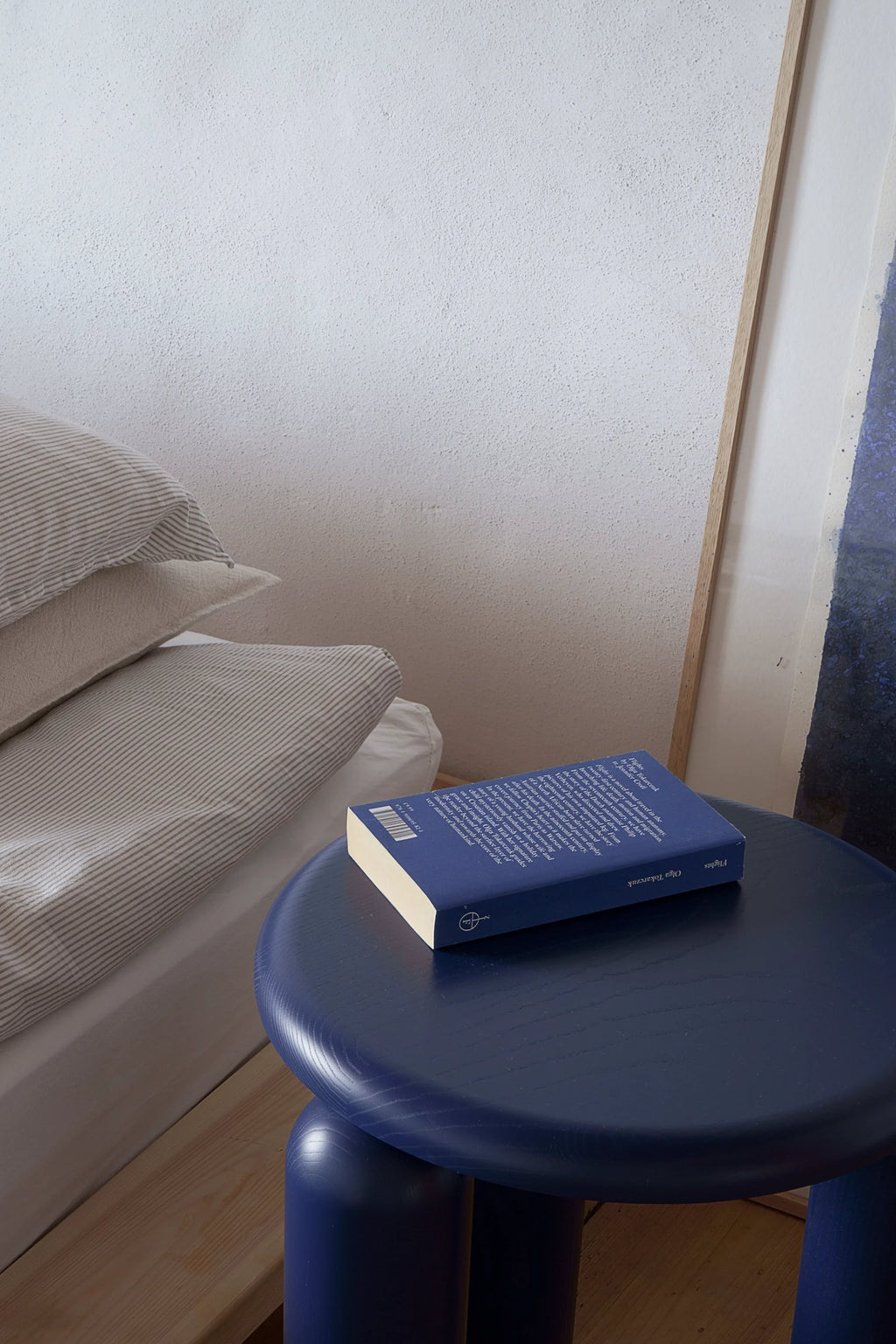 TAMO Neo Side Table in a lifestyle setting, showcasing its rich, deep navy blue lacquered wood finish with visible grain and distinctive rounded tripod legs. A book rests on its minimalist top, beside a bed with striped pillows. This premium furniture piece is perfect for modern bedrooms and living spaces.