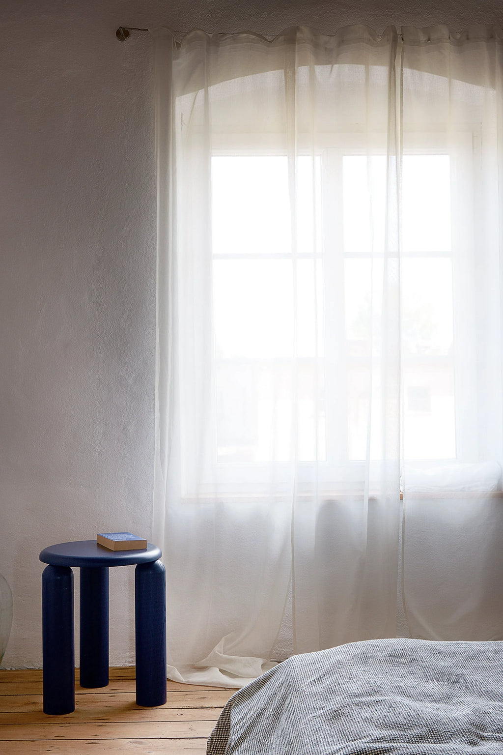 TAMO Neo Side Table in a natural setting. The modern navy blue cylindrical stool with a flat round top sits on a rustic wooden floor next to a bed with a grey textured blanket and sheer white curtains. Ideal for contemporary living spaces.