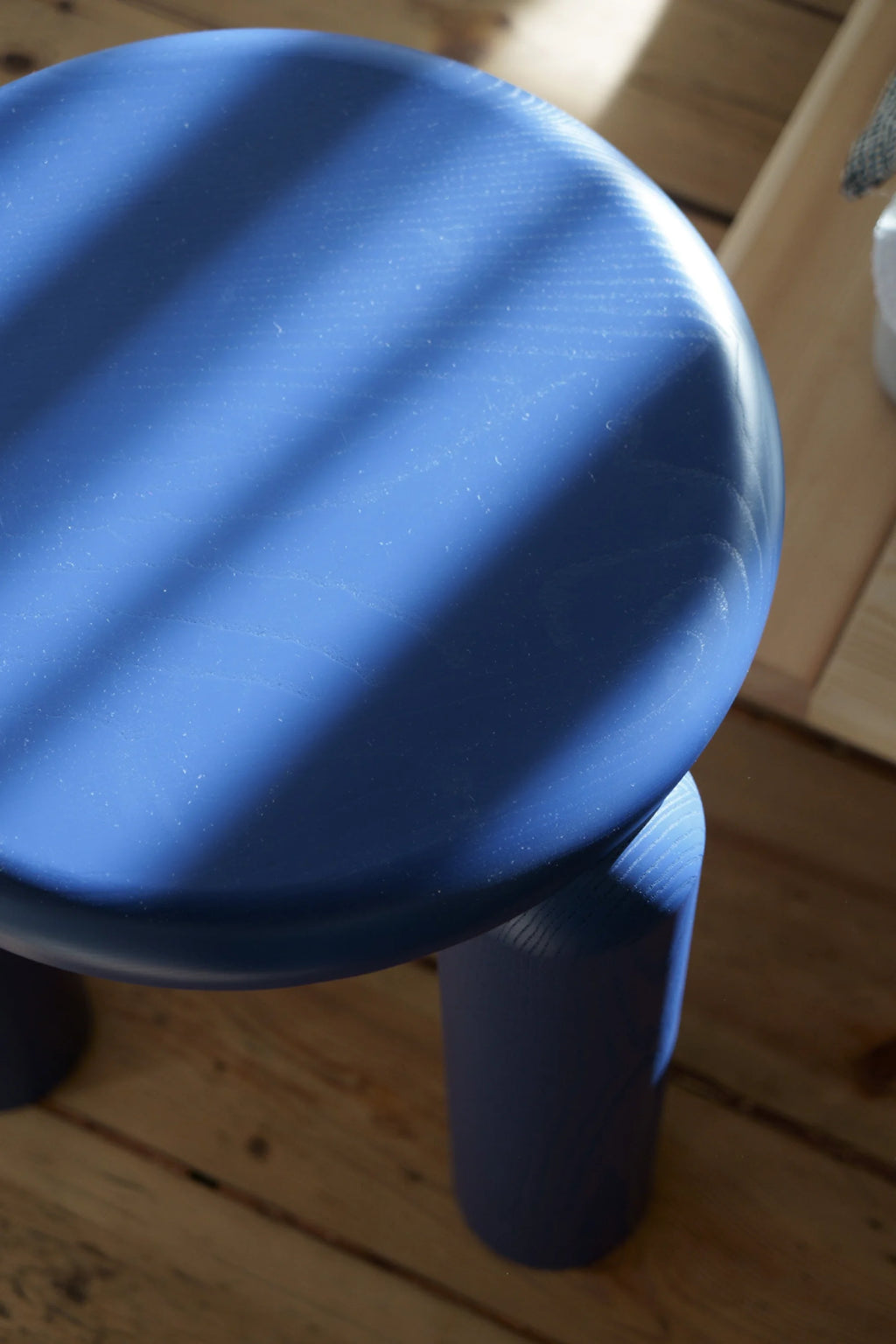 TAMO Neo Side Table in a detailed close-up, showing its round, vibrant royal blue wooden top and one thick cylindrical leg. Light highlights the visible grain of this premium contemporary furniture, placed on a warm wooden floor. Ideal for modern living spaces.