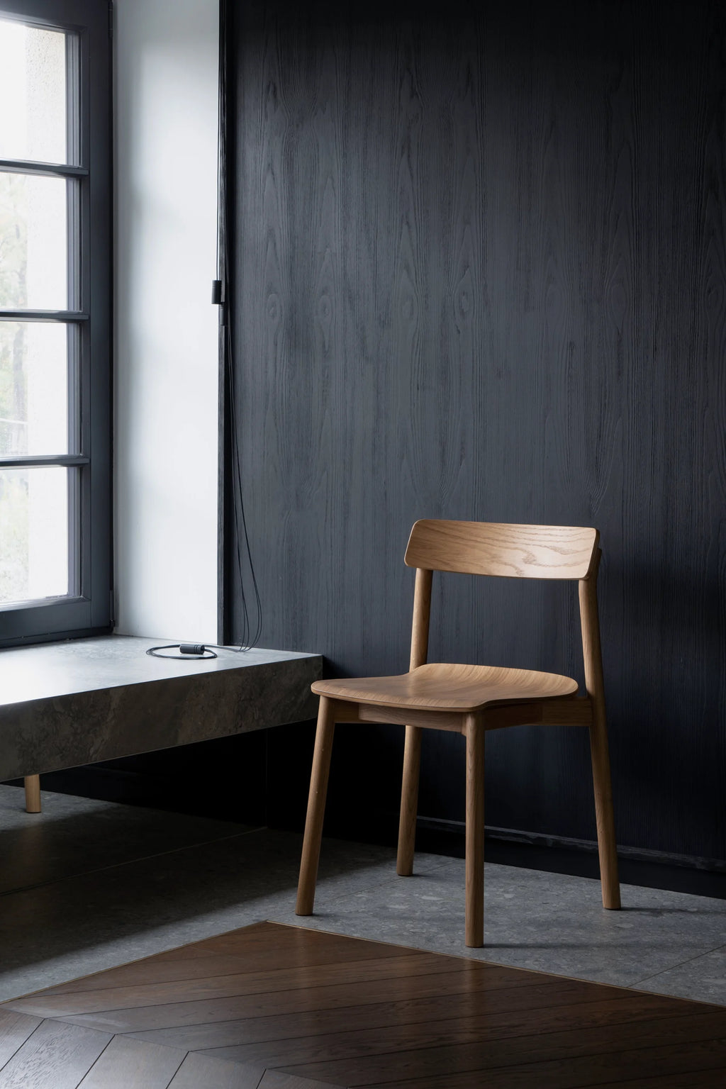 TAMO Arche Chair in a lifestyle shot, showcasing its curved light brown solid wood seat and back, and four tapered legs, against a dark wooden wall. This authentic wooden dining chair is perfect for modern interiors.