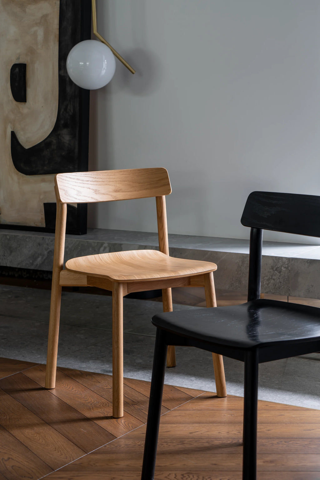 TAMO Arche Chair. Two modern dining chairs, one light natural wood and one matte black, with subtly curved backs and seats, displayed on a rich wood floor. Ideal for contemporary interior design.