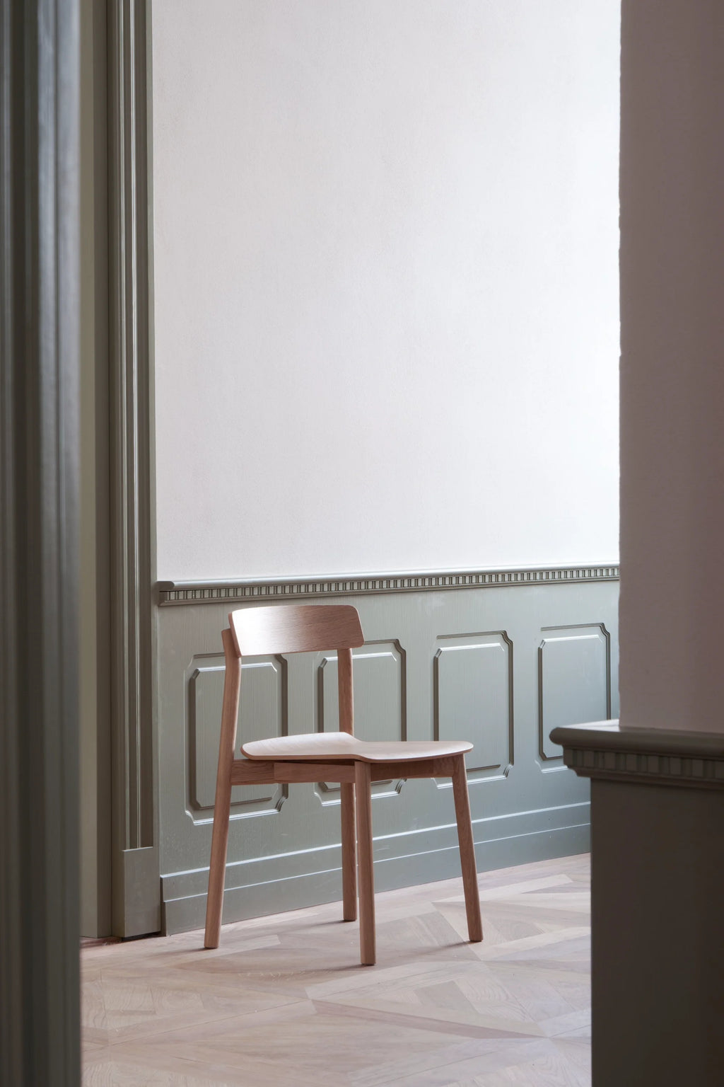 TAMO Arche Chair, a premium light oak wood dining chair, featuring a curved backrest & seat, displayed on light parquet flooring against sage green wainscoting. Ideal for modern living spaces.