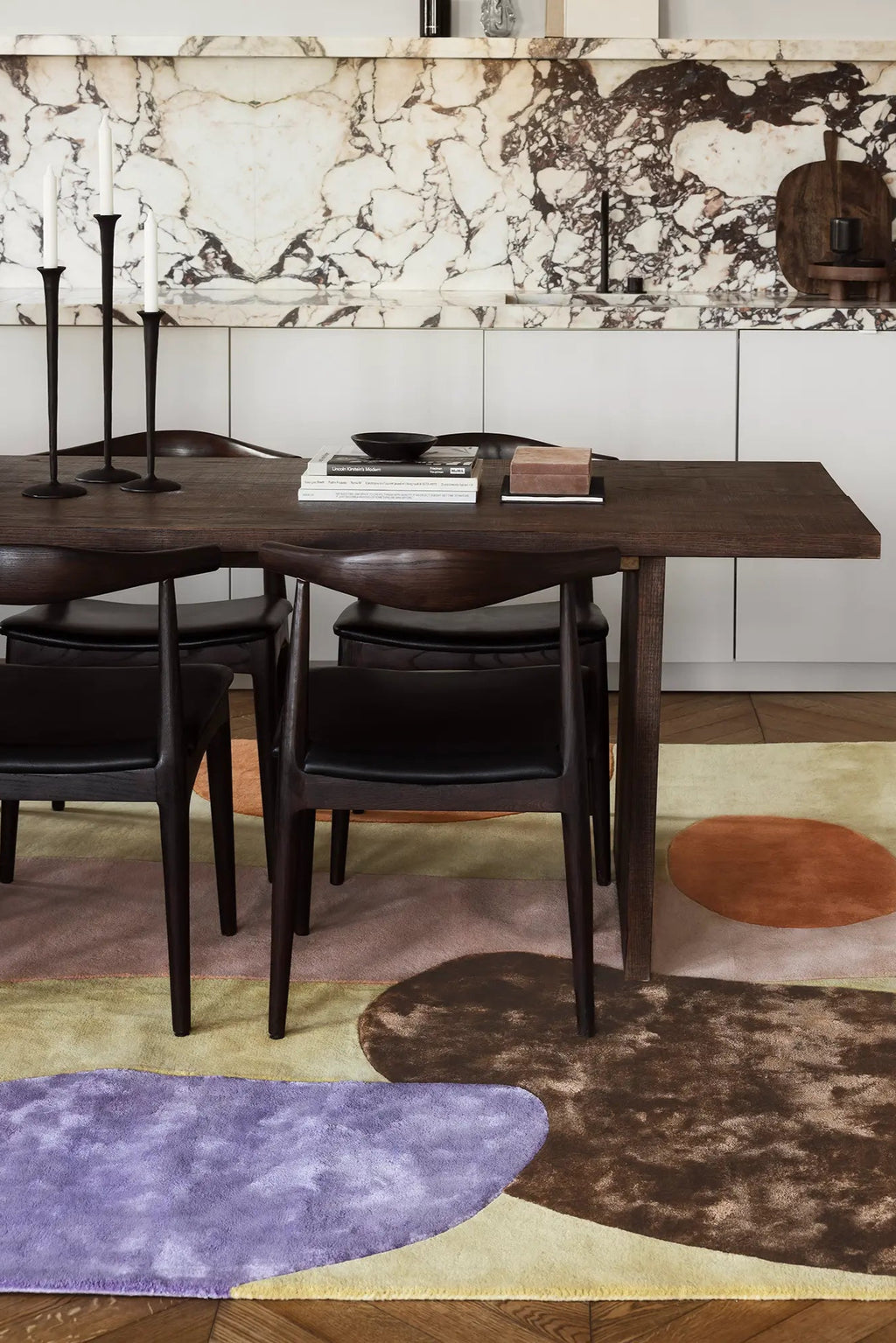 Layered Magical Garden Wool Rug, featuring an abstract, colorful design with organic shapes in shades of purple, brown, orange, and pale yellow, visible under a dark wood dining table. Perfect for...