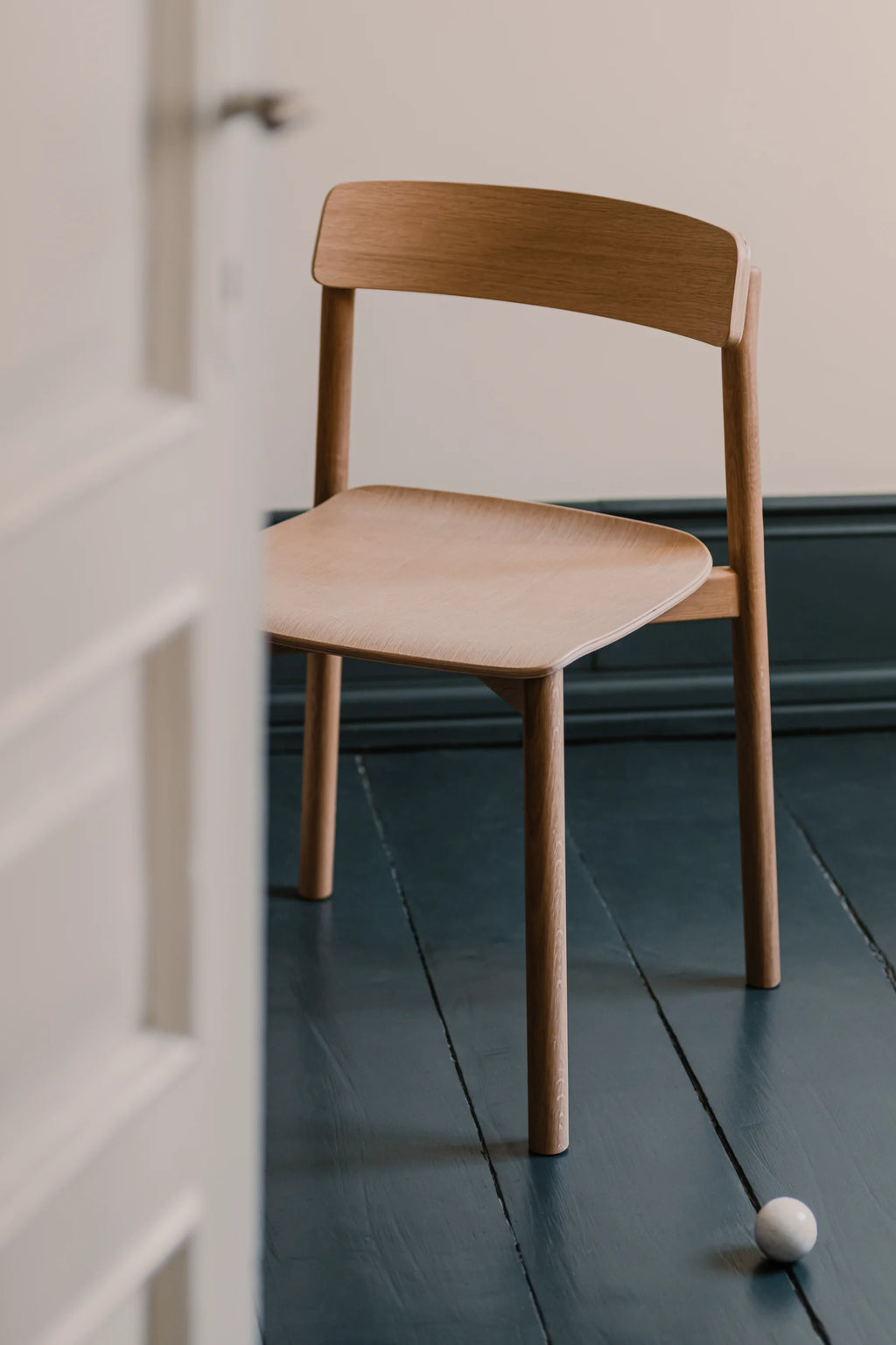 TAMO Arche Chair, a premium light oak wood dining chair with a curved backrest and square seat, presented on a dark blue wooden floor. Ideal for modern minimalist interiors.