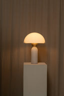 Table lamp with a beige shade on a white pedestal against a brown curtain background