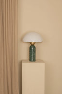 Table lamp with green base and white shade on a beige pedestal against a beige wall.
