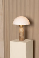 Table lamp with a white shade and bronze base on a beige pedestal against a beige curtain.
