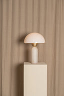 Minimalist table lamp with a white shade and gold base on a beige pedestal against a beige curtain.