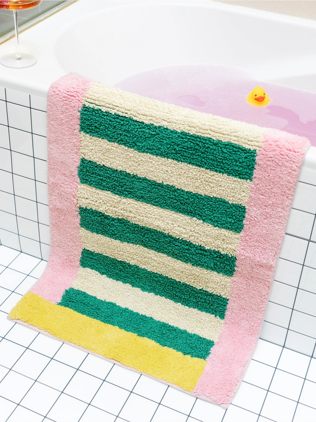 TARTA GELATINA Piña Colada Bath Mat, a vibrant and playful retro bath mat crafted from plush, absorbent tufted cotton with a shaggy texture. It features bold horizontal stripes in cream, emerald green, and a sunny yellow base, all framed by a cheerful candy pink border, creating a striking rectangular design. This colorful striped bath mat is ideal for adding a fun, maximalist touch to a modern bathroom or a kids' bathroom decor.