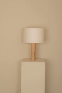 Table lamp with wooden base and white shade on a beige pedestal against a beige wall.