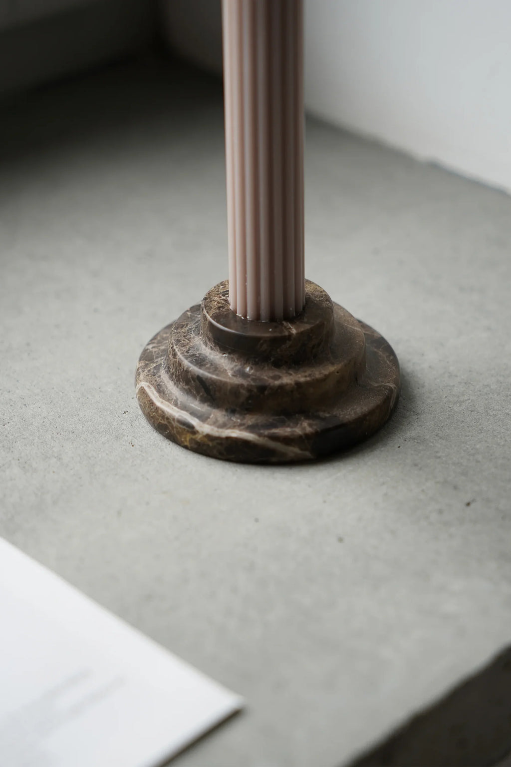 TAMO Rokk Candle Holder with a pale pink fluted candle set in a dark brown polished marble base, featuring natural white and amber veining, displayed on a light gray concrete surface. A luxury home decor accent.