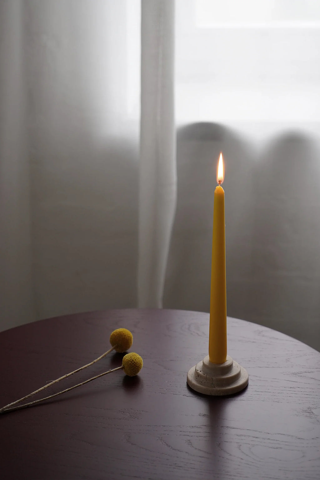 TAMO Rokk Candle Holder: A premium, minimalist off-white circular stone holder with stepped edges, displaying a lit yellow taper candle on a dark wood table. Perfect for atmospheric home decor.