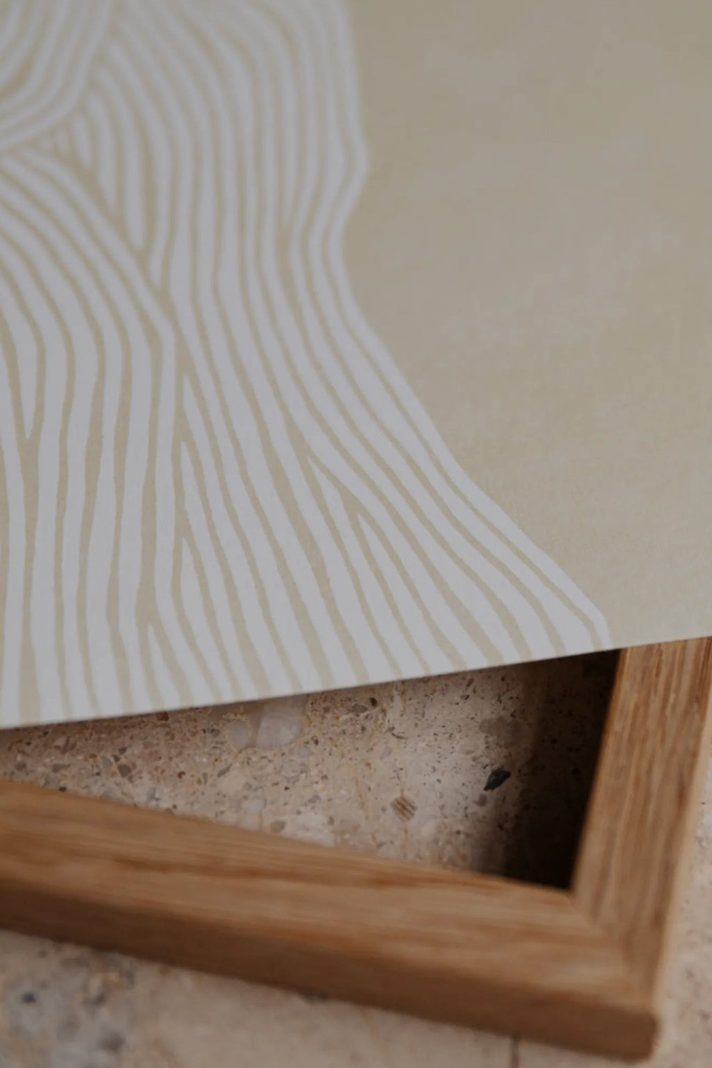 The Poster Club The Line no. 07, a minimalist abstract art print featuring organic off-white wavy lines on a soft beige background, creating a subtle, textured visual effect. This contemporary paper art print is partially shown with a warm natural oak frame. Ideal for adding a serene touch to Scandinavian living rooms, organic modern bedrooms, or elegant gallery walls.