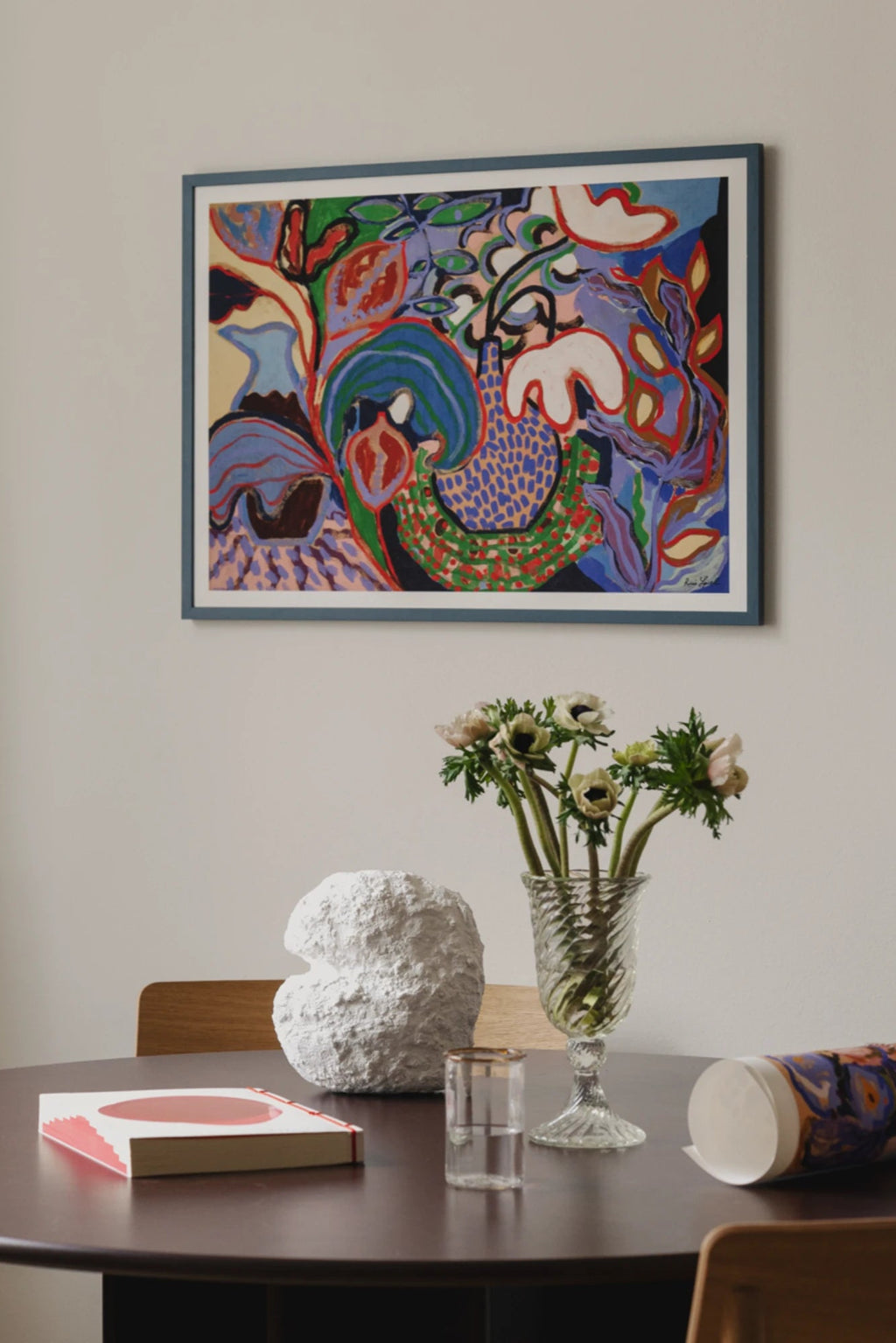 The Poster Club Felicity, a vibrant abstract art print featuring a rich tapestry of organic shapes and bold brushstrokes in blues, purples, reds, greens, and yellows, framed in a sleek muted blue frame. This colorful modern wall art is perfect for adding a dynamic focal point to contemporary living rooms or dining spaces.