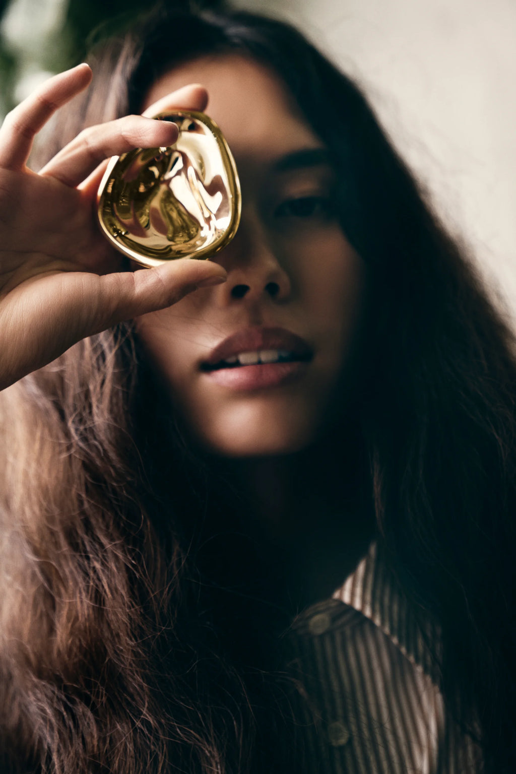 Hein Studio Ripple Hook/Knob: Close-up of a model's hand holding a luxurious gold-finished, organically shaped ripple hook/knob over her eye, demonstrating its unique form. Perfect for adding modern, artistic flair.