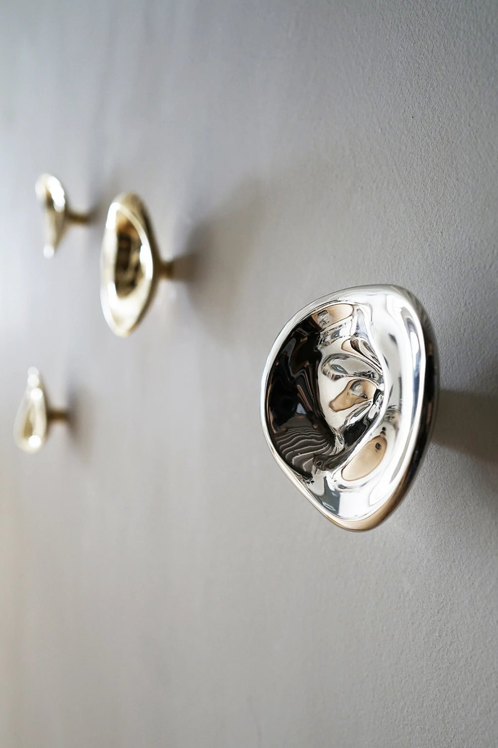 Hein Studio Ripple Hook/Knob in a close-up, depicting two gold and one prominent silver organic-shaped, glossy, premium brass hooks mounted on a textured grey wall. Ideal for contemporary interior design.