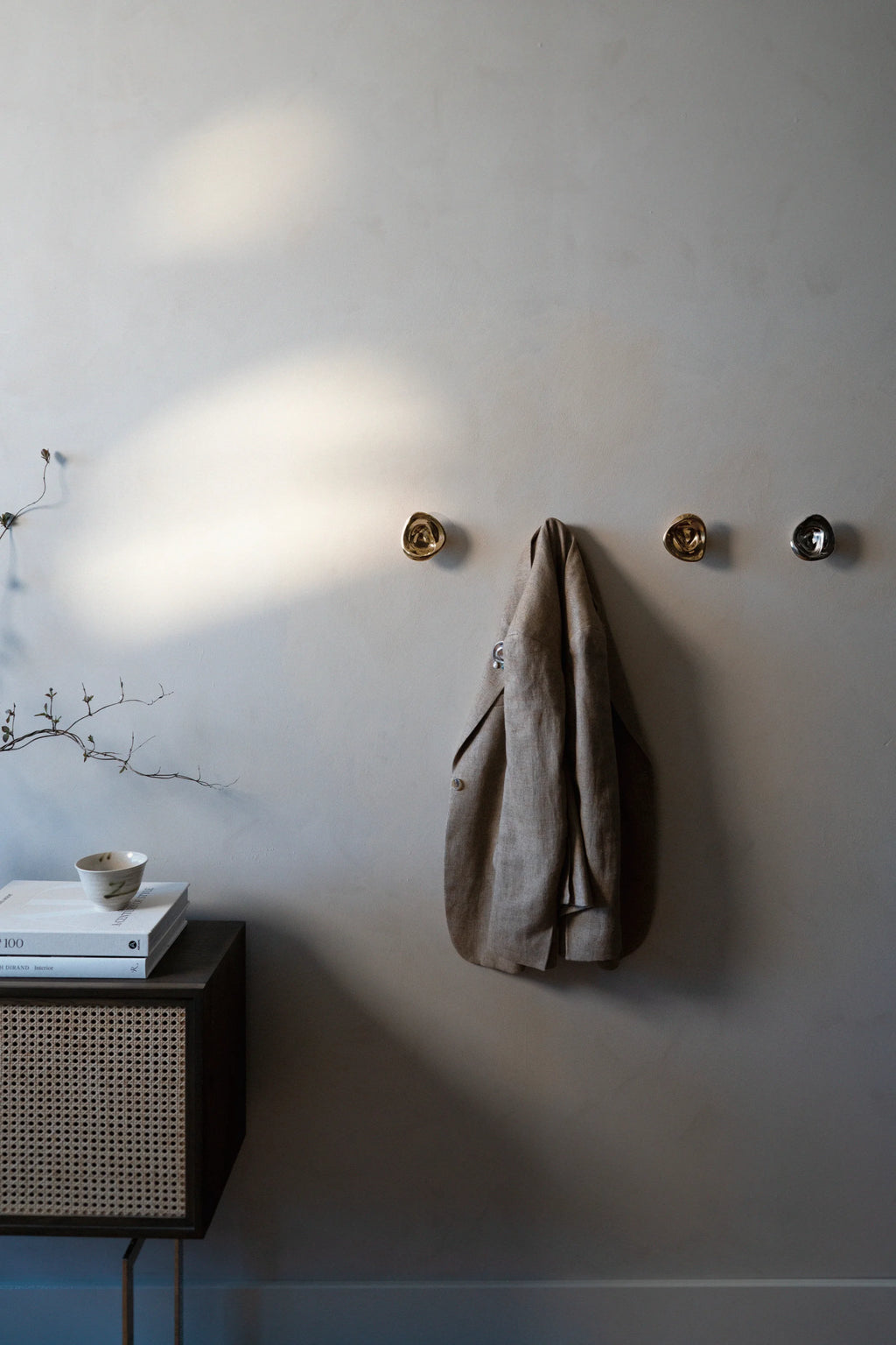 Hein Studio Ripple Hook/Knob in a lifestyle shot: three luxury brass-gold and dark gray ripple-textured metal wall hooks/knobs mounted on a light gray wall, one holding a beige linen jacket. Ideal for contemporary interior design.