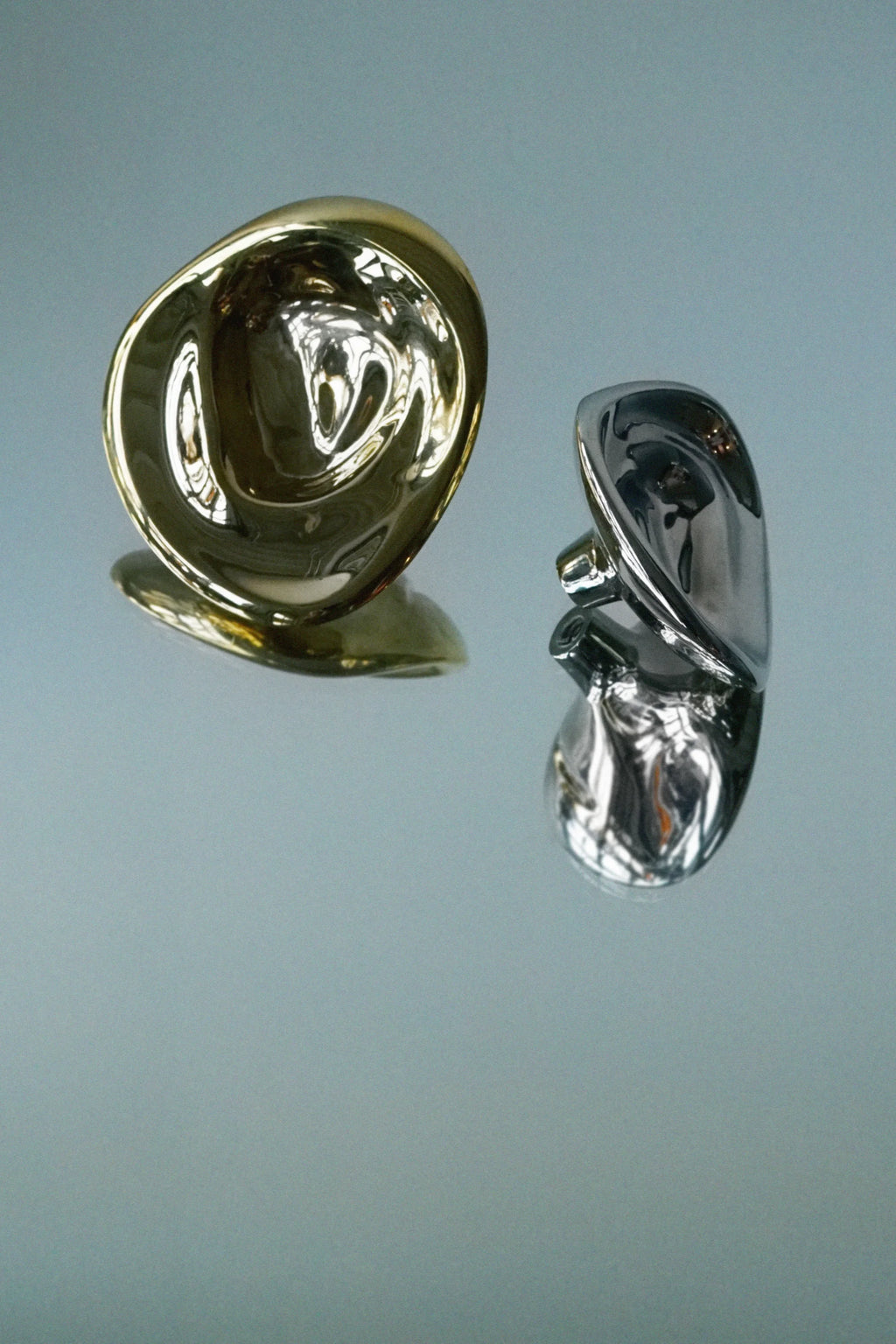 Hein Studio's Ripple Hook/Knob set, featuring one shiny gold and one silver organic-shaped, luxury metal knob, displayed with reflections on a light blue-grey surface. Ideal for modern interior design.