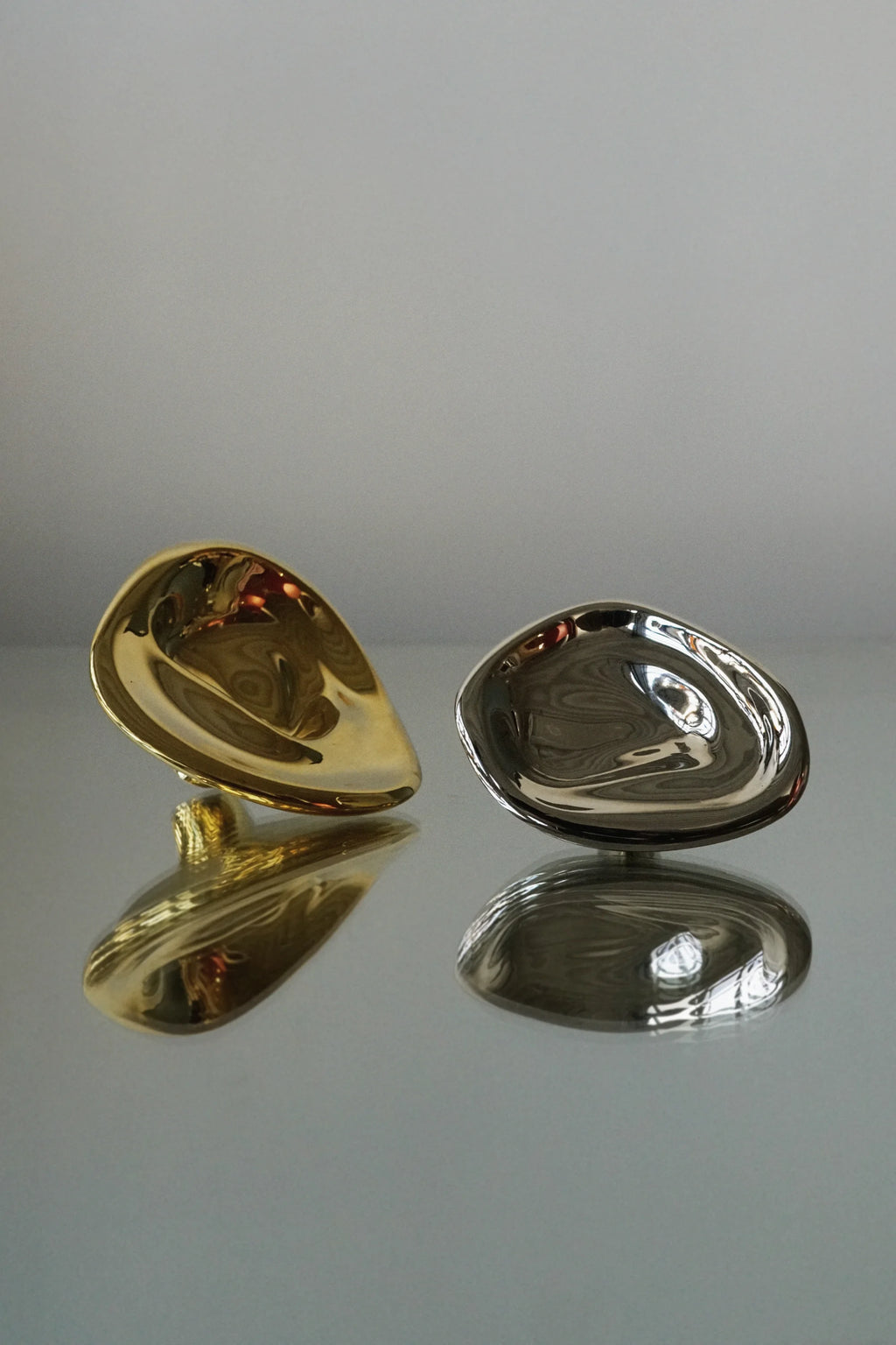 Hein Studio Ripple Hook/Knob: Two luxury teardrop-shaped metal hooks, one gleaming golden and one polished silver, shown from an elevated angle with reflections on a glossy surface. Perfect for contemporary interior design.