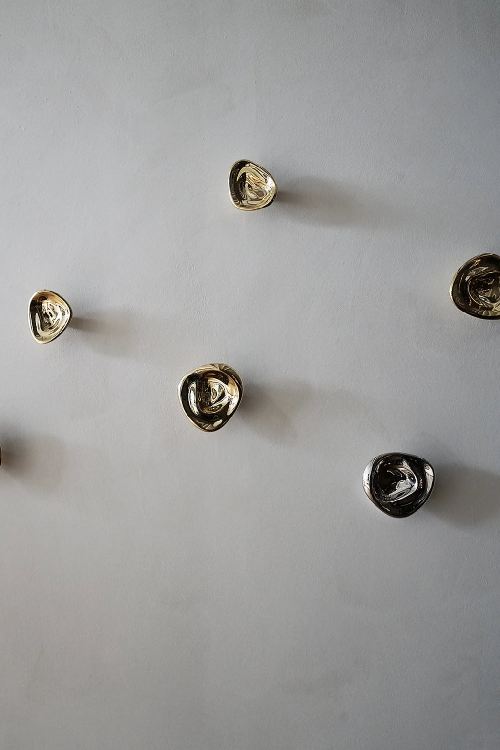 Hein Studio's Ripple Hooks/Knobs, a luxurious collection of seven metallic, organically shaped wall hooks in both gold and silver finishes, arranged artistically on a light gray wall. Perfect for modern interior decor.