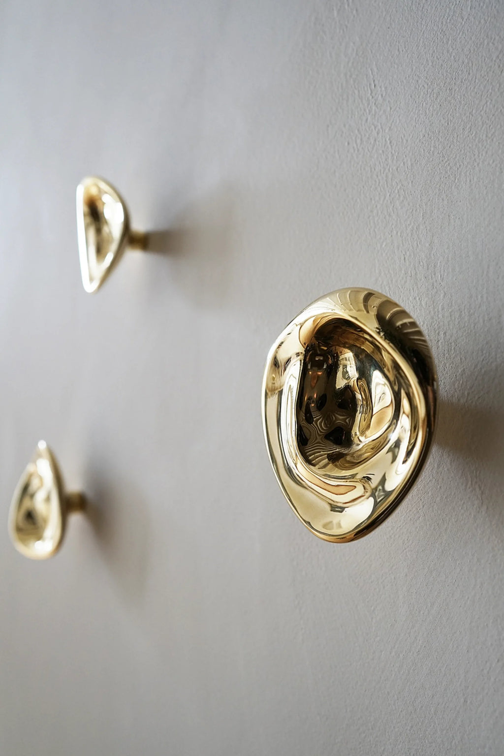Hein Studio Ripple Hook/Knob: Three luxurious, reflective gold-toned irregular-shaped hooks with a hammered, organic texture mounted on a light gray wall. A premium decorative and functional accent.