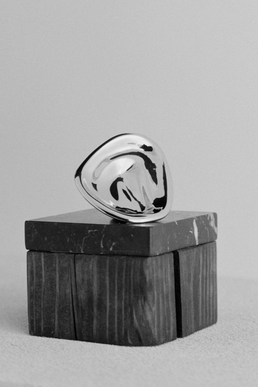 Hein Studio Ripple Hook/Knob in a highly polished, organic, fluid silver-toned metal, displayed on a textured dark marble block, against a light gray background. This luxury furniture hardware is perfect for adding designer appeal.