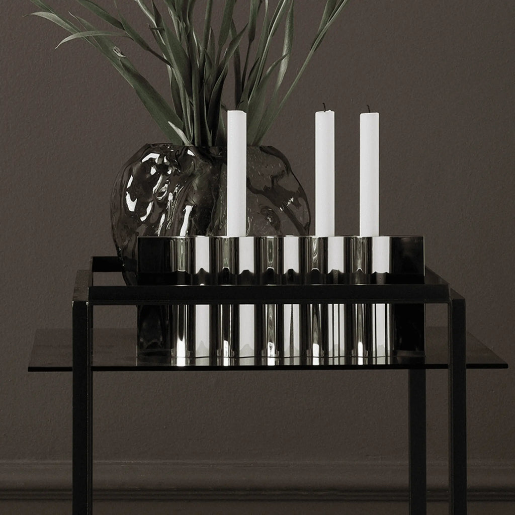 New Works Ripply Candle Holder, a modern sculptural candle holder crafted from highly polished silver-toned stainless steel with a distinctive fluted and ribbed surface that creates dynamic reflections. This contemporary metal candle holder features multiple slots for white taper candles, offering a minimalist yet impactful design. It's an ideal table centerpiece for modern dining rooms or a sophisticated accent for living room decor.