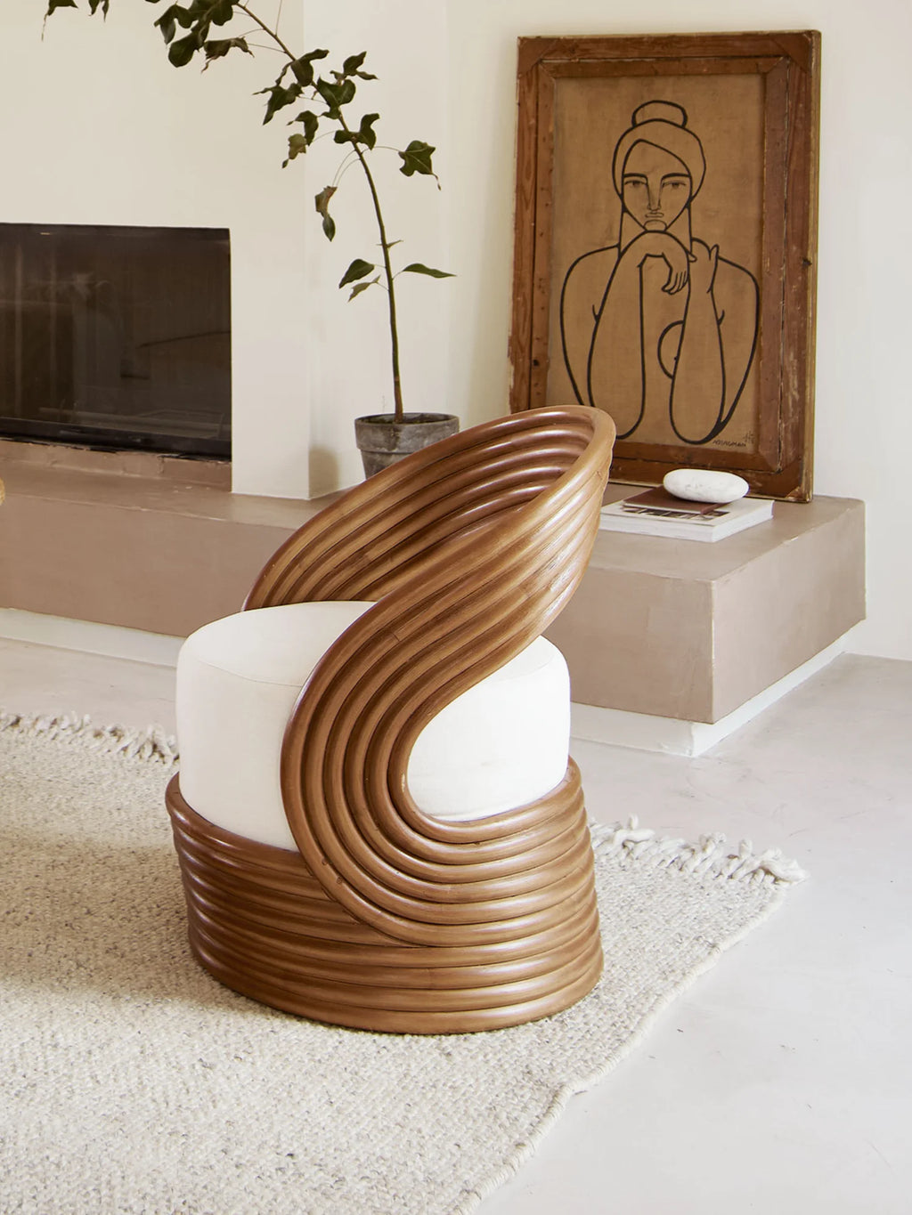 Tine K Mano Armchair, a sculptural organic modern lounge chair crafted from natural light brown curved rattan, forming a stacked, undulating silhouette that embraces a plush round off-white upholstered seat. This unique accent chair is perfect for contemporary living rooms, cozy reading nooks, or coastal-inspired bedrooms, adding a touch of natural elegance.