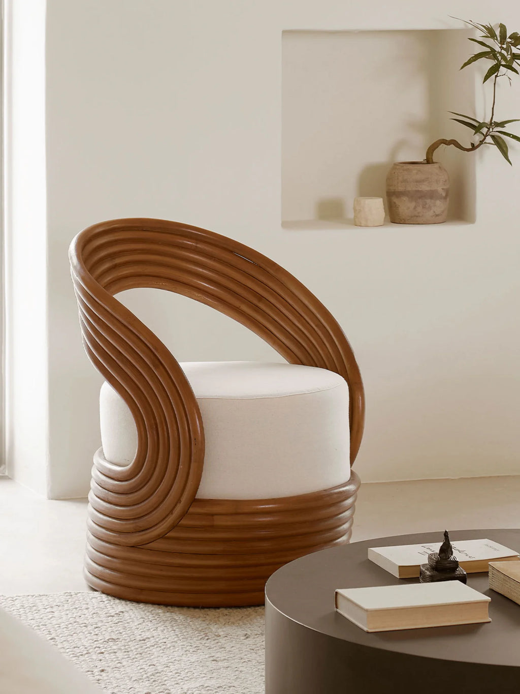Tine K Mano Armchair, a sculptural organic modern lounge chair featuring a unique spiraled natural rattan frame in warm brown tones, wrapping around a plush cylindrical cream upholstered seat. This contemporary accent chair showcases a distinctive coiled rattan design forming both the backrest and base, offering a striking visual texture and comfortable seating. Ideal as a statement piece for boho living rooms, coastal bedrooms, or a stylish reading nook.