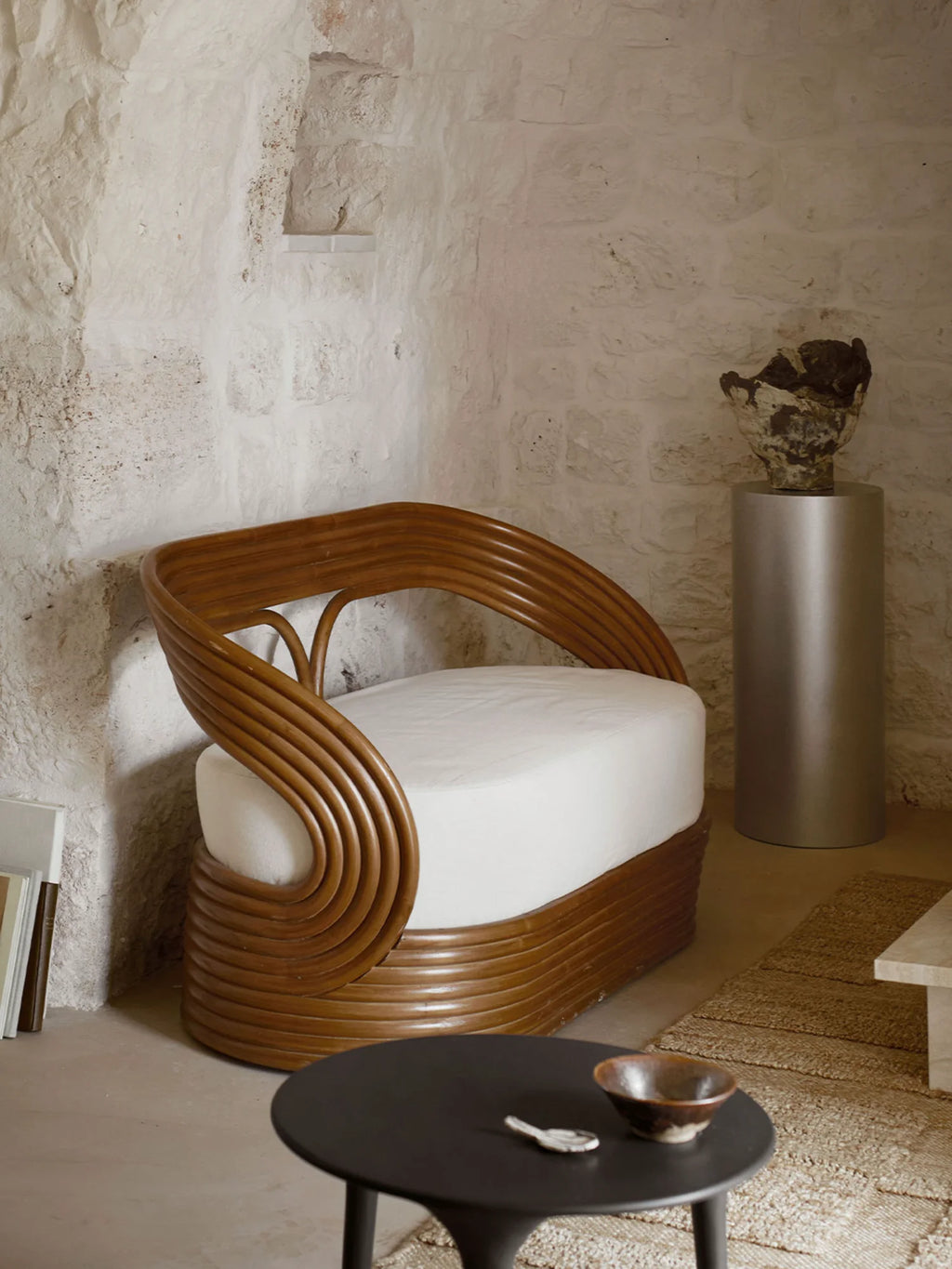 Tine K Mano Sofa, an organic modern lounge sofa crafted with a distinctive curved natural rattan frame featuring stacked, hand-bent rattan strips in a warm brown finish, complemented by a plush cream.