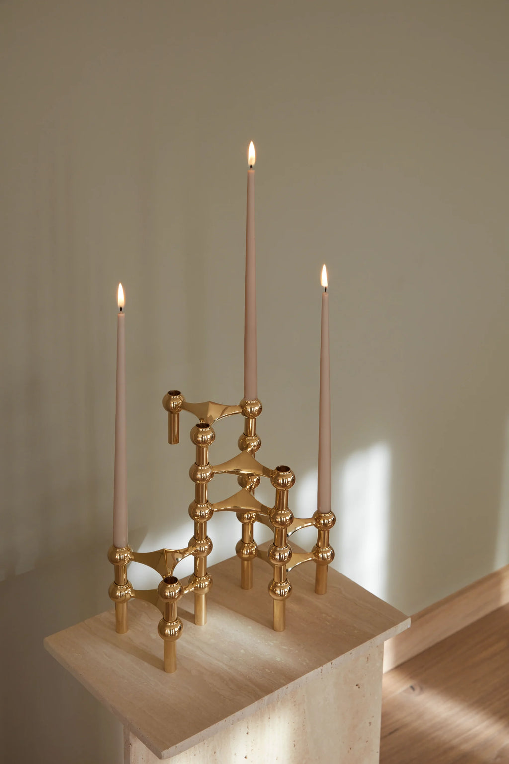 Stoff Nagel Candle Holder in a radiant polished brass finish, configured into an asymmetrical, artistic display with three lit beige taper candles, resting on a textured light stone surface.
