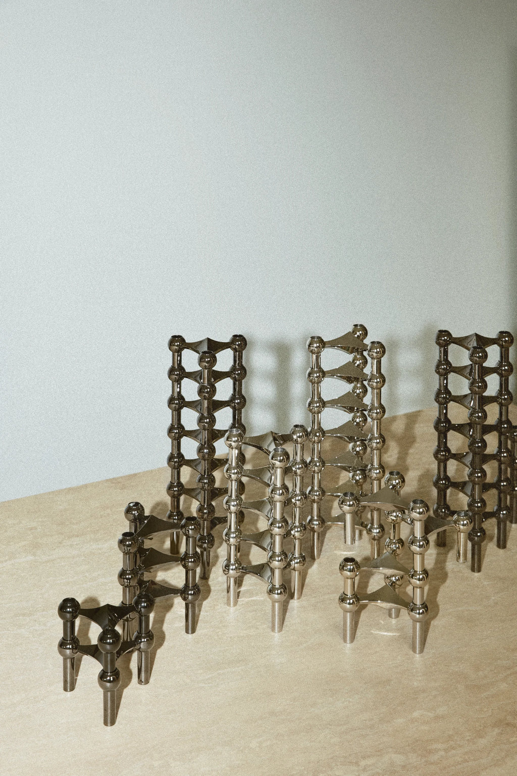Stoff Nagel Candle Holder, a modular, sculptural design in polished silver-toned metal, featuring multiple interconnected spherical and rod elements, arranged on a light beige marble surface against a soft gray wall, showcasing its elegant, minimalist aesthetic.