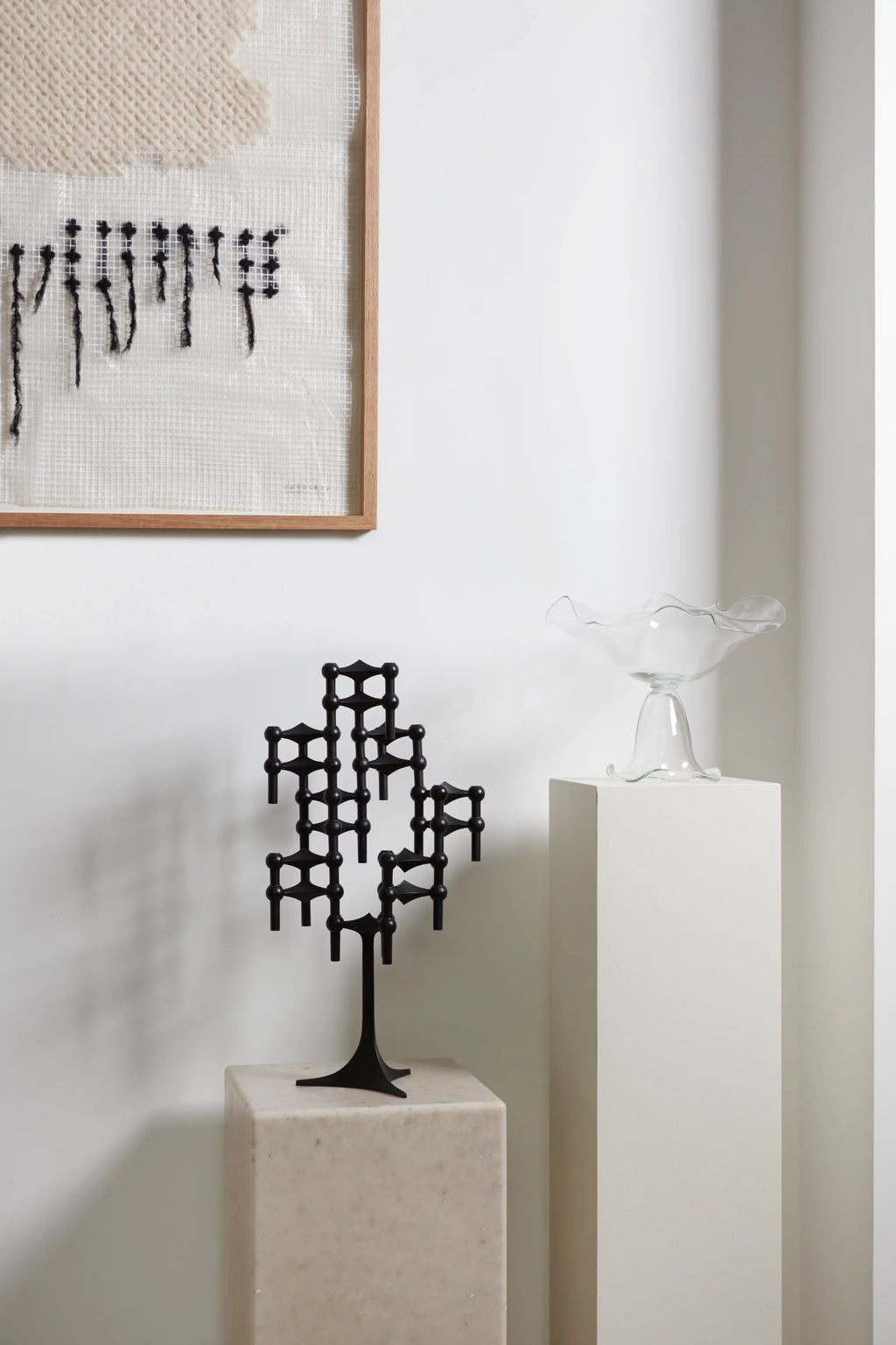 Stoff Nagel Candle Holder in a sophisticated matte black finish, assembled into a captivating, abstract sculpture, placed on a light-colored stone pedestal in a bright, modern interior.