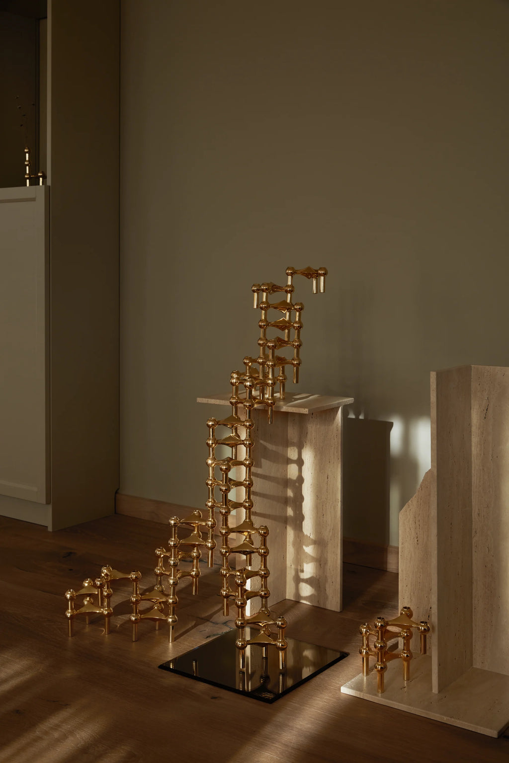 Multiple Stoff Nagel Candle Holders in a vibrant brass finish, arranged in various configurations, including a tall, spiraling sculpture and smaller clusters, displayed on a wooden floor with travertine pedestals.