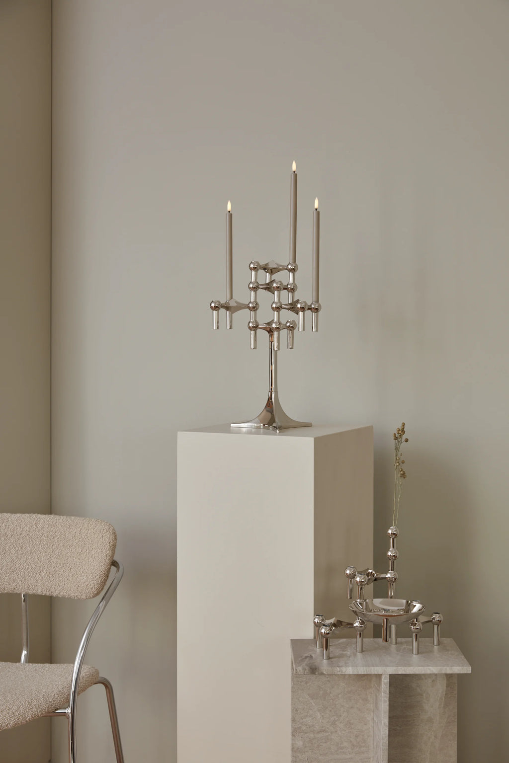 Stoff Nagel Candle Holder in a gleaming chrome finish, assembled into a tall, elegant candelabra holding three lit grey taper candles, displayed on a clean white pedestal in a modern interior.