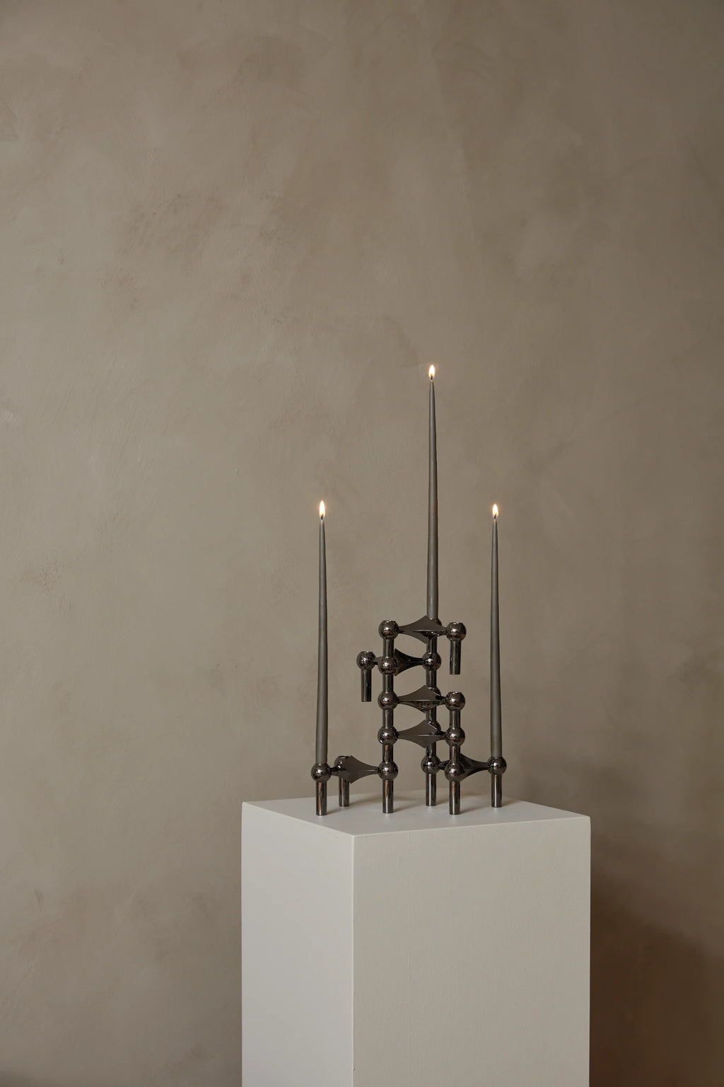 Stoff Nagel Candle Holder in a dark, gunmetal finish, forming a compact, intricate structure with three lit charcoal-gray taper candles, set on a crisp white pedestal against a textured wall.