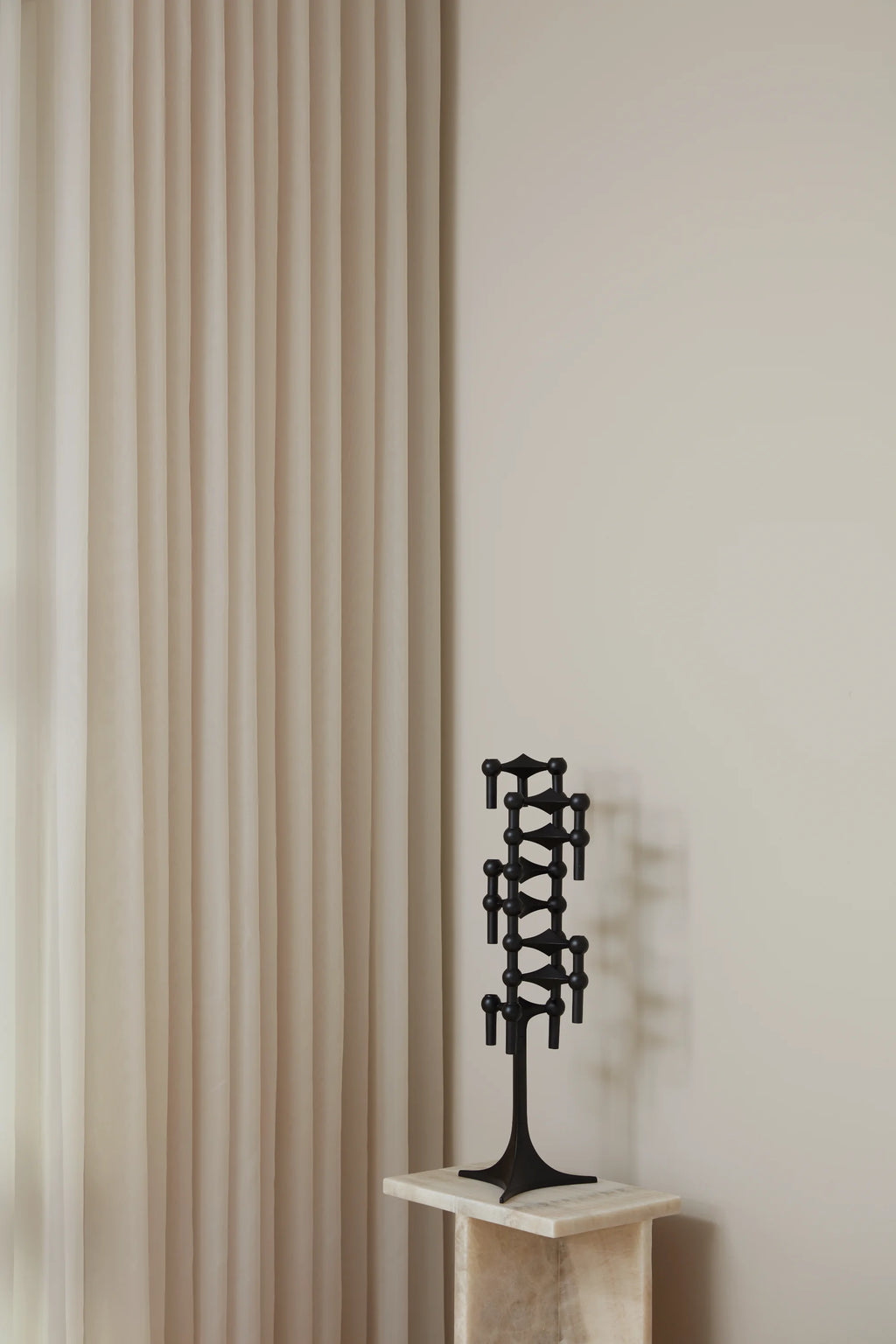 Stoff Nagel Candle Holder in a striking matte black finish, assembled into a tall, elegant, multi-tiered candelabra, positioned on a light marble pedestal next to flowing sheer curtains.