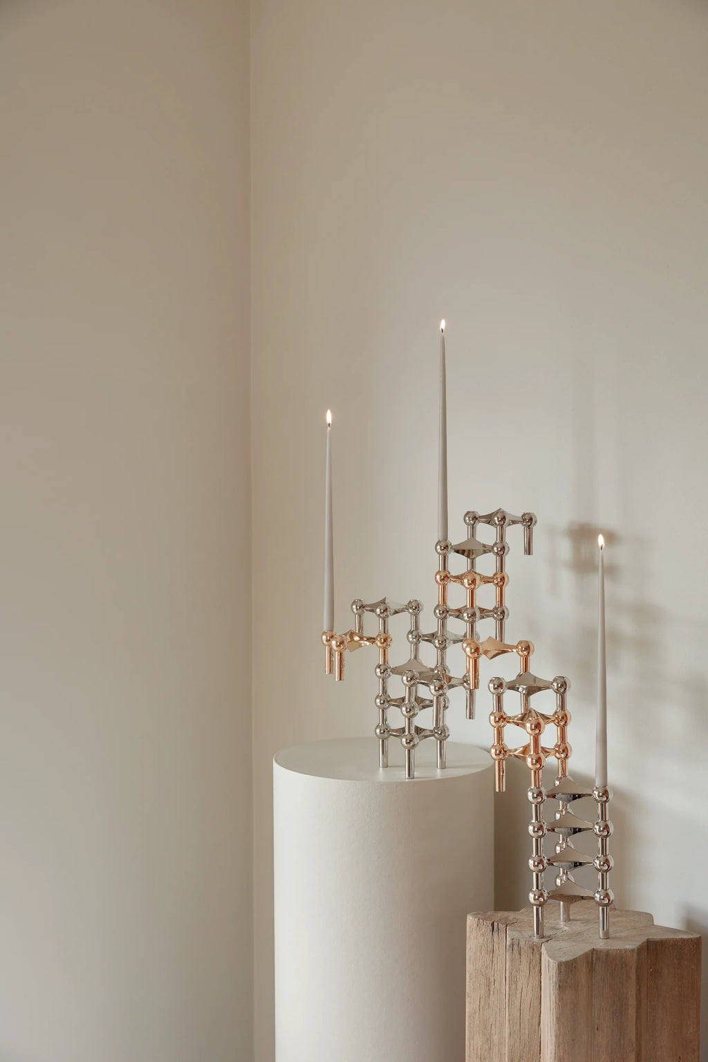 Stoff Nagel Candle Holder collection featuring a mix of chrome and copper finishes, forming an elegant, multi-layered display with three lit gray taper candles on a white cylindrical pedestal.