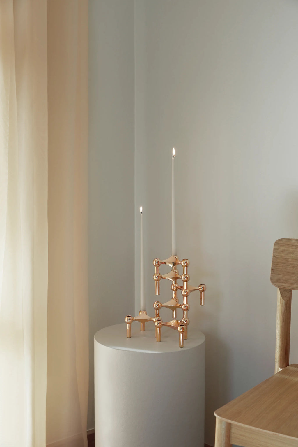 Stoff Nagel Candle Holder in a warm, polished copper finish, arranged with two lit white taper candles, creating a cozy, inviting glow on a round white pedestal in a sunlit room.