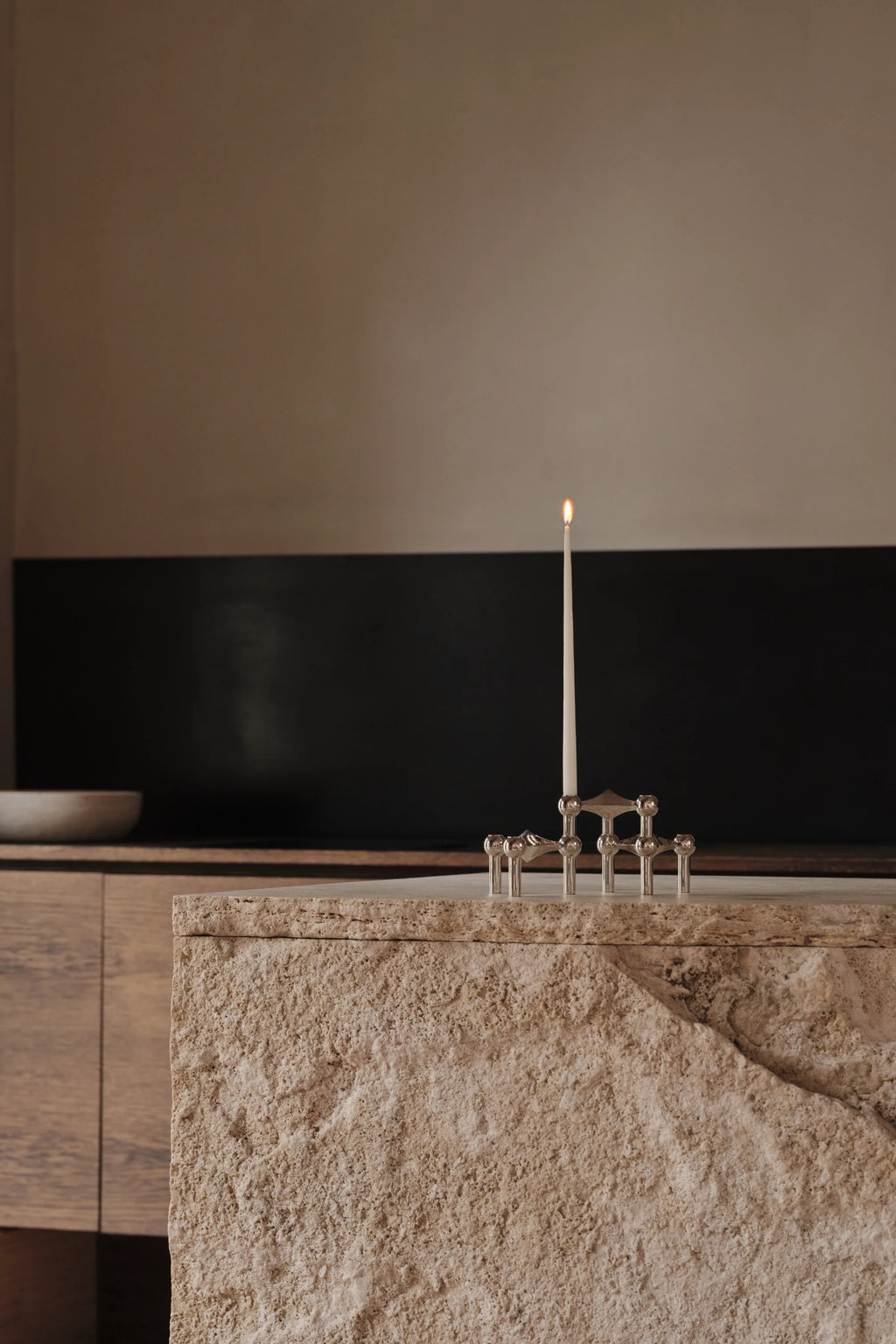 Stoff Nagel Candle Holder in a polished chrome finish, configured to hold a single lit cream taper candle, elegantly positioned on a rough-hewn, light-colored stone counter in a contemporary kitchen setting.