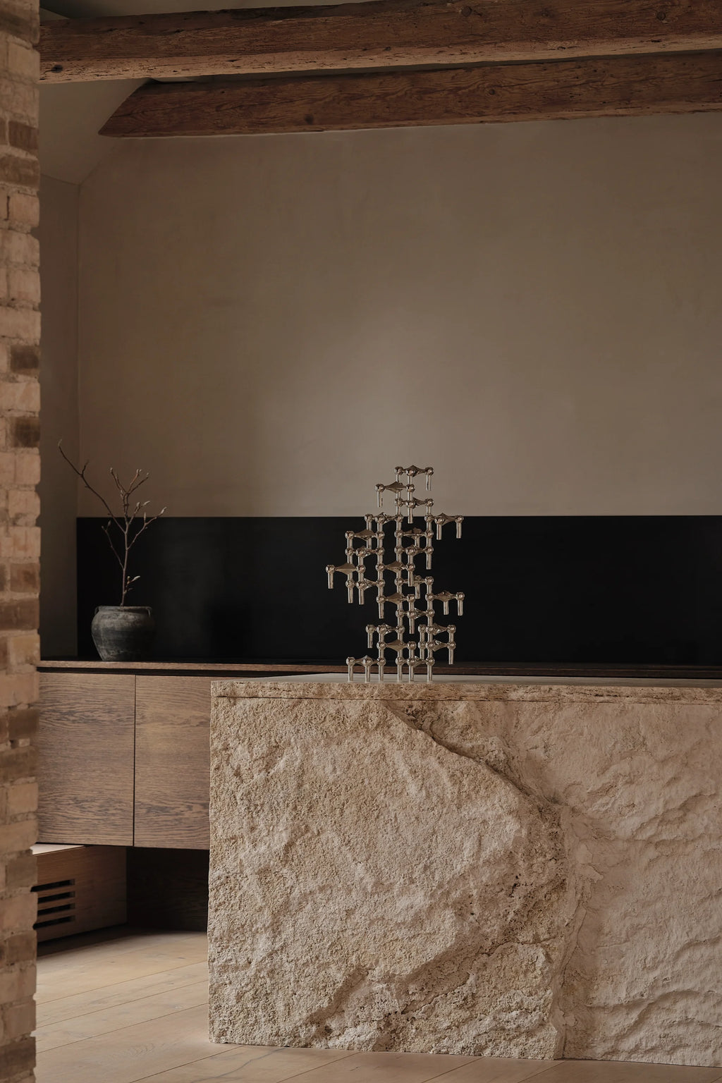 Stoff Nagel Candle Holder in a polished chrome finish, intricately built into a complex, multi-tiered sculpture, prominently displayed on a large, textured stone island in a modern, rustic-inspired kitchen.