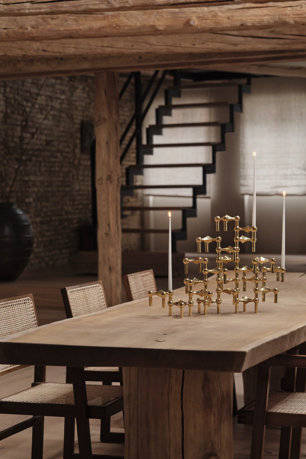 Stoff Nagel Candle Holder in a radiant brass finish, intricately assembled into a large, impressive centerpiece holding three lit white taper candles, gracing a rustic wooden dining table with cane chairs.