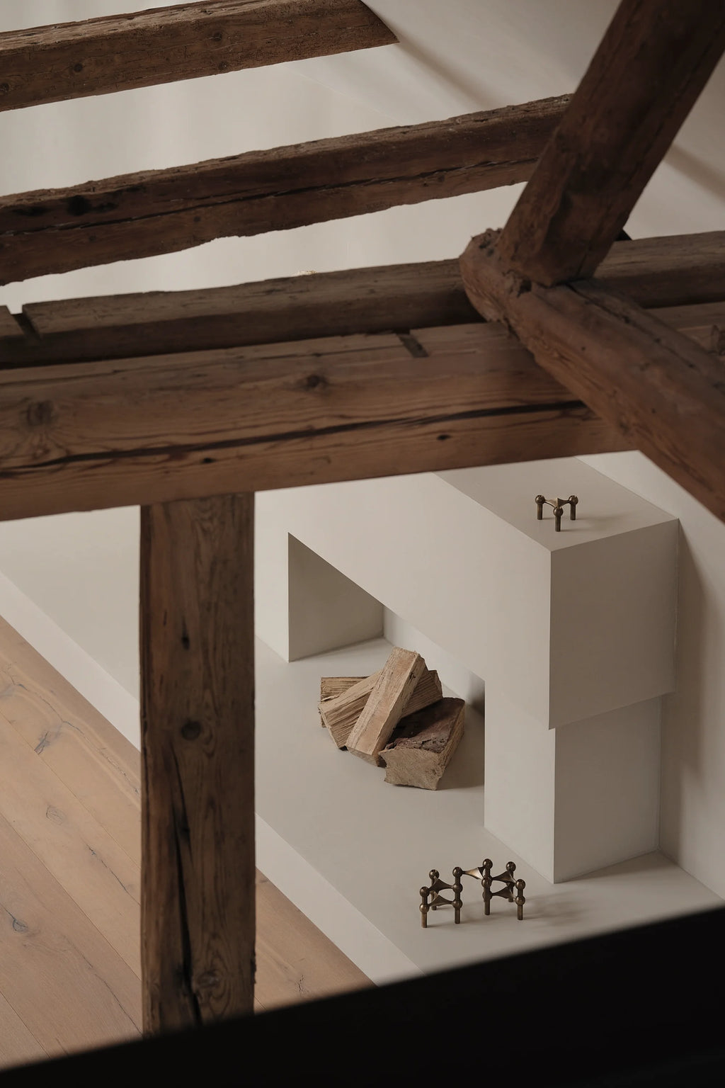 Two Stoff Nagel Candle Holders in a warm brass finish, one small and one larger, positioned on a minimalist white fireplace mantel with stacked logs, viewed from above with rustic wooden beams.