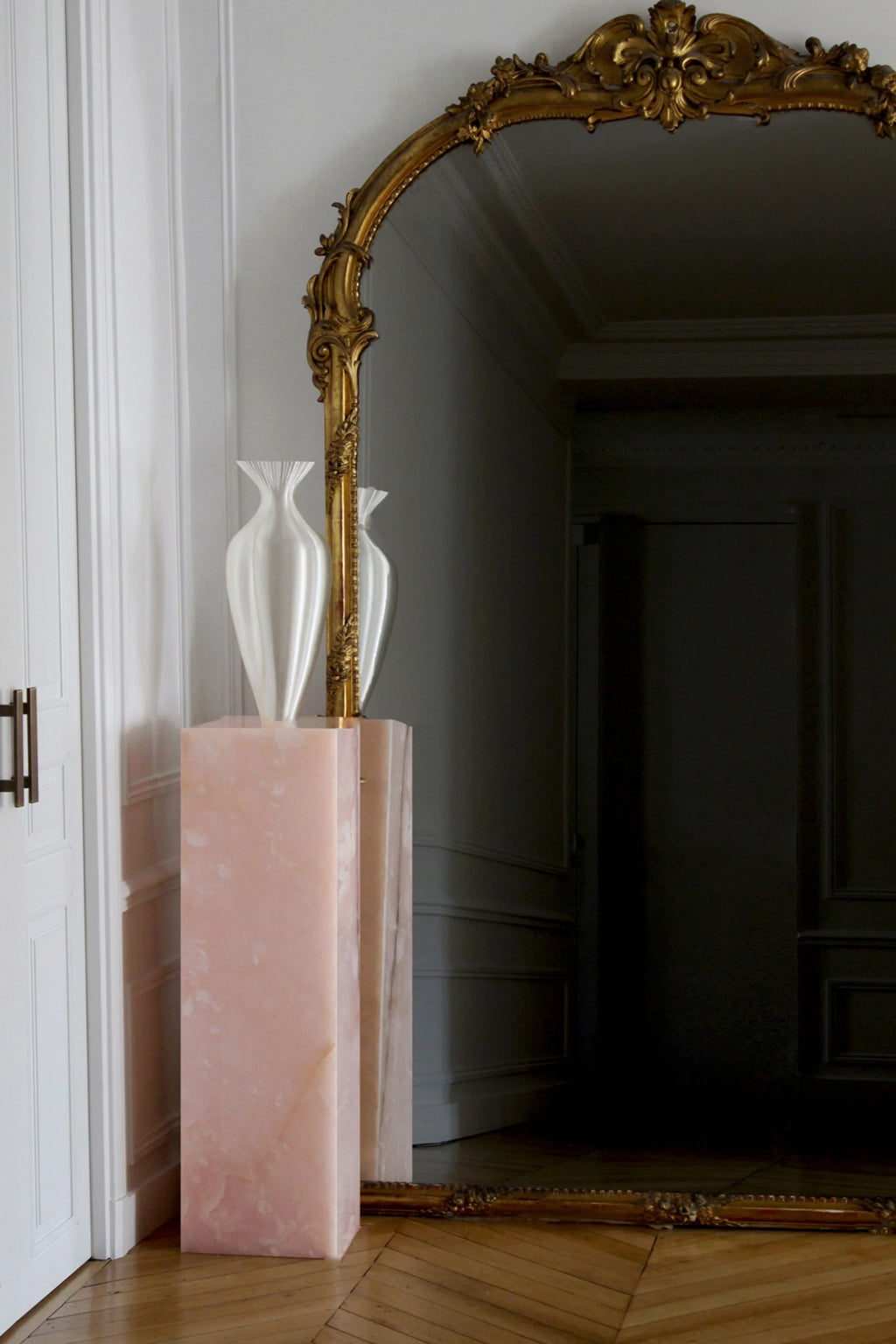 Decorative vase with white flowers on a pink pedestal in front of an ornate gold mirror.