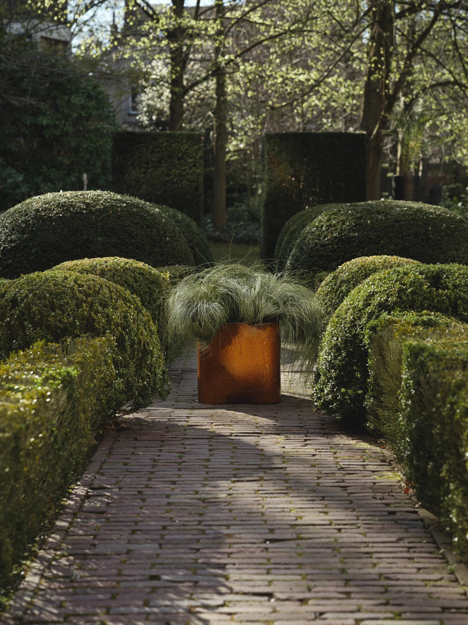A square, ceramic Book Planter in a glossy, deeply mottled burnt orange/caramel glaze is centered on a brick pathway that recedes into a garden. The planter is filled with a soft, mounding green ornamental grass. The pathway is framed by low, neatly clipped boxwood hedges and large, rounded topiaries, with taller, square topiaries visible in the distance. The scene is outdoors on a sunny day.