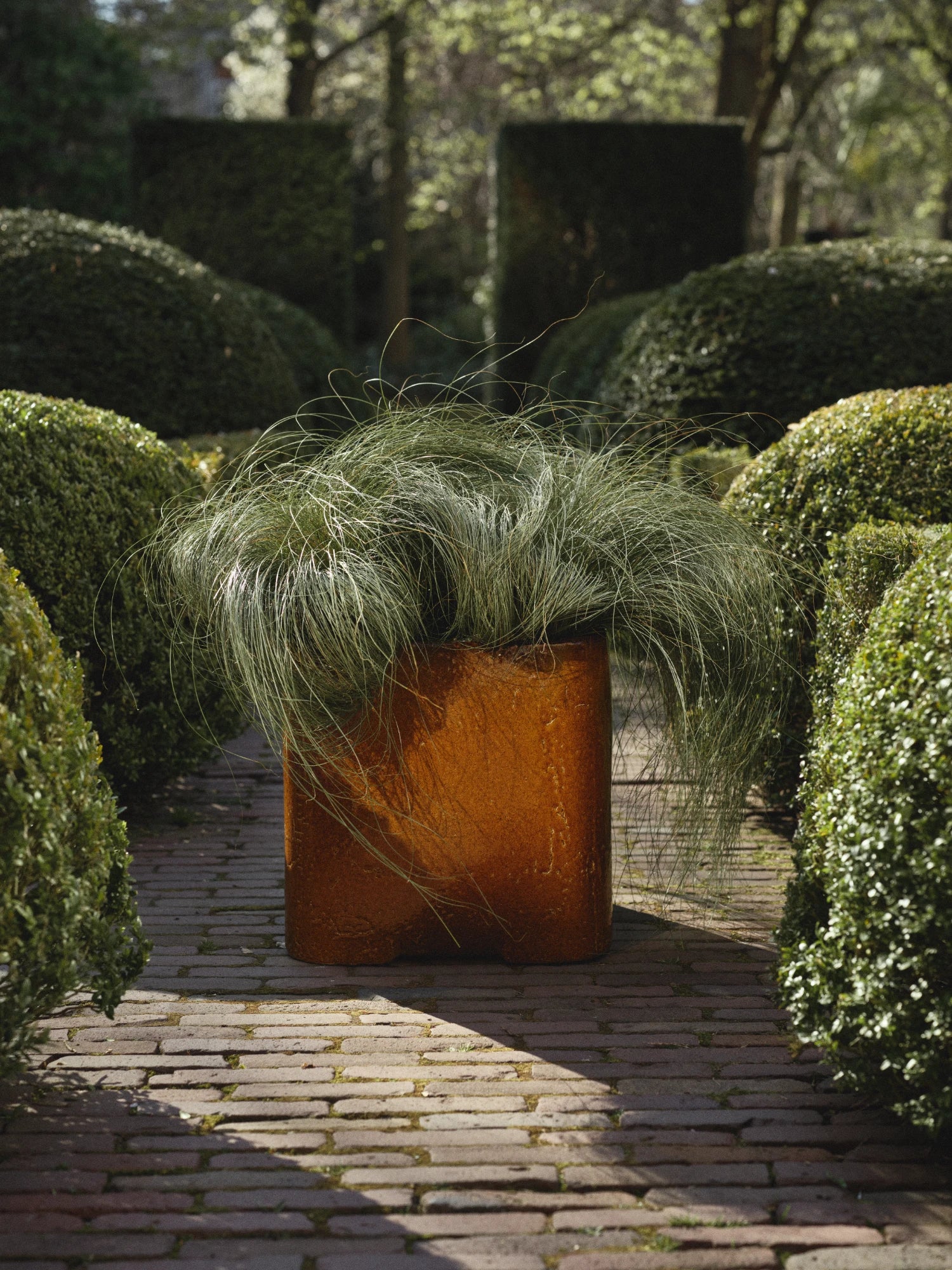 A tall, cylindrical ceramic Book Planter 04 in a glossy, deeply mottled burnt orange/caramel glaze is placed on a brick pathway in a formal garden. The planter is filled with a cascading, fine-bladed green ornamental grass. The background is a lush, manicured garden with sculpted boxwood hedges and topiary forms.
